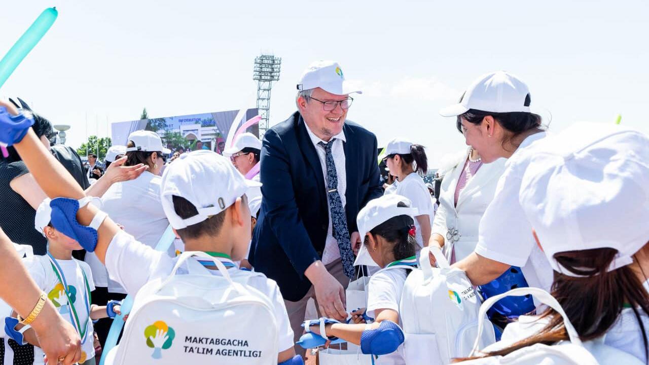 OSCE Project Co-ordinator in Uzbekistan interacts with a group of children in white uniforms and caps during an outdoor event.